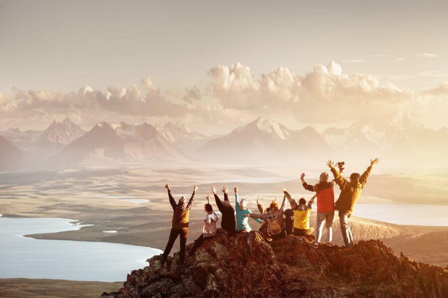 Big group of people having fun in success pose with raised arms on mountain top against sunset lakes and mountains